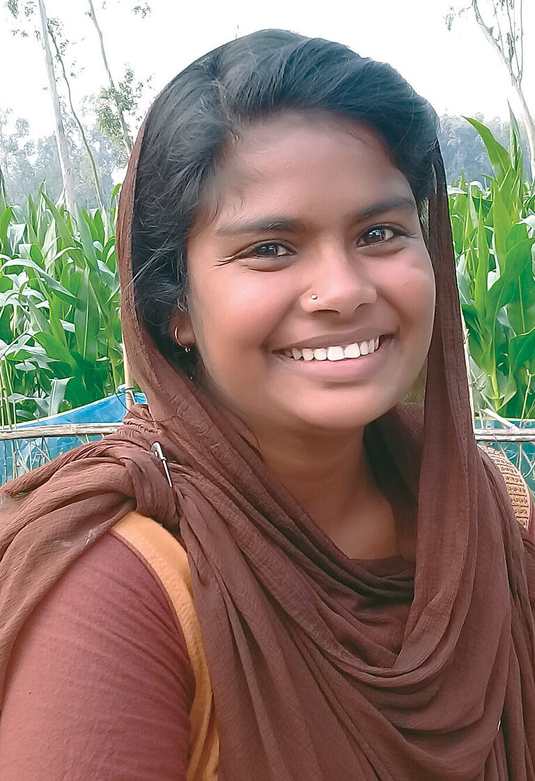 Ein Mädchen steht vor einem Maisfeld und schaut lachend in die Kamera.