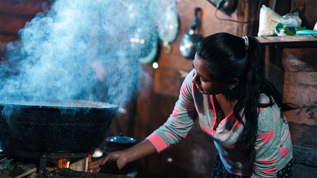 Eine Frau legt Holz auf das Feuer in ihrer Küche. Über dem Feuer dampft ein großer Kochtopf.