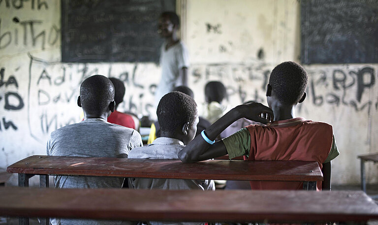 Auf einer hölzernen Schulbank sitzen in einem dunklen Klassenzimmer drei Jungen in T-Shirts nebeneinander und blicken zur Tafel und dem Lehrer
