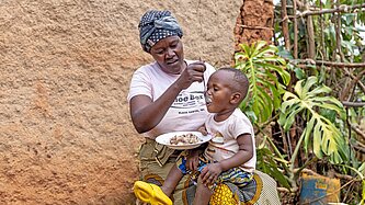 Ein kleiner Junge wird von seiner Mutter gefüttert, die vor einem Lehmhaus sitzt