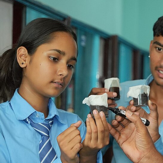 Ein Mädchen und ein Junge aus Nepal, beide in Schuluniform, analysieren eine Flüssigkeit in einem Messbecher