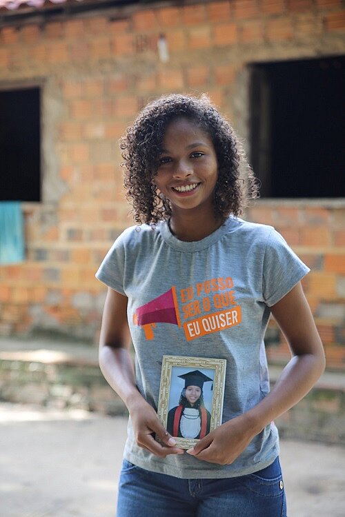 Patenkind Cassia Eine junge Frau steht im T-Shirt vor einem Gebäude, sie hält ein Bild von sich beim Schulabschluss in der Hand