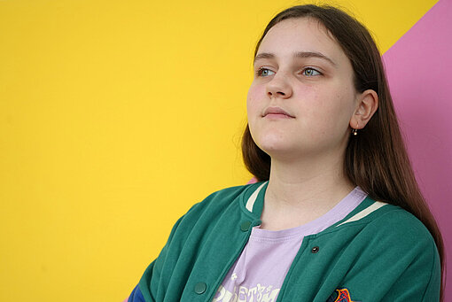 Eine Teenagerin steht vor einer bunt angemalten Wand und schaut in die Ferne. Sie hat braune Haare und trägt eine grüne Sportjacke.