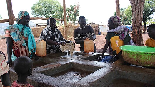 Frauen und Kinder stehen um eine Waschstation und holen sich unterhaltend Wasser bzw. waschen ihre Kleidung.