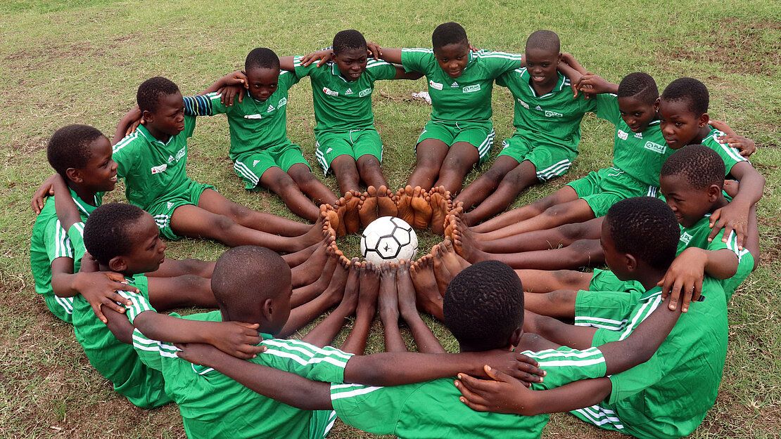 Eine Gruppe Kinder in grünen Trikots sitzt um einen Fußball herum auf den Boden, sie haben ihre Arme umeinander gelegt. 