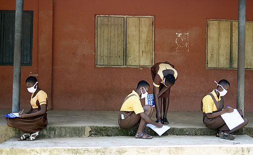 Plan International setzt sich dafür ein, dass der Stellenwert von Bildung, insbesondere für Mädchen, in den Fokus gerückt wird. Vier Mädchen sitzen vor dem Schulgebäude und erledigen Aufgaben.