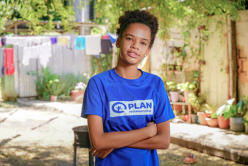 Melany nimmt an Plan-Projekten teil Ein Mädchen trägt ein Plan-T-Shirt, hat die Arme vor der Brust verschränkt und schaut in die Kamera