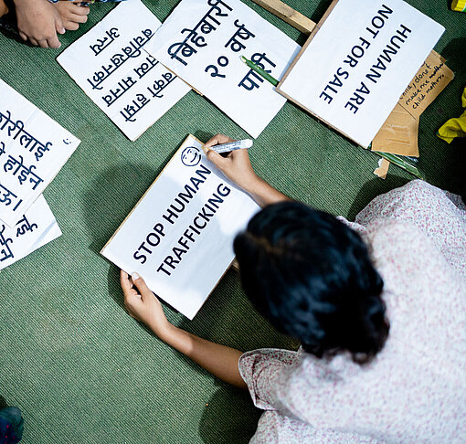 Auf einem grünen Boden liegen unterschiedliche Schilder mit nepalesischer Schrift. Darüber gebeugt sitzen junge Frauen