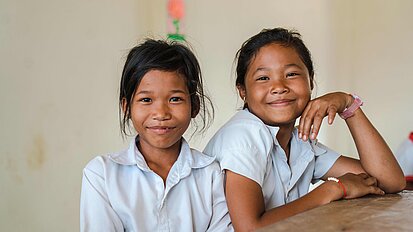 Zwei Mädchen aus Kambodscha sitzen in ihrer Schule an einem Tisch und lächeln in die Kamera.