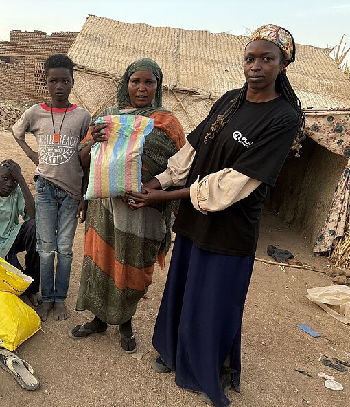 Eine Helferin in Plan-Weste übergibt Lebensmittelhilfen an Frauen und Kinder in einem Camp.