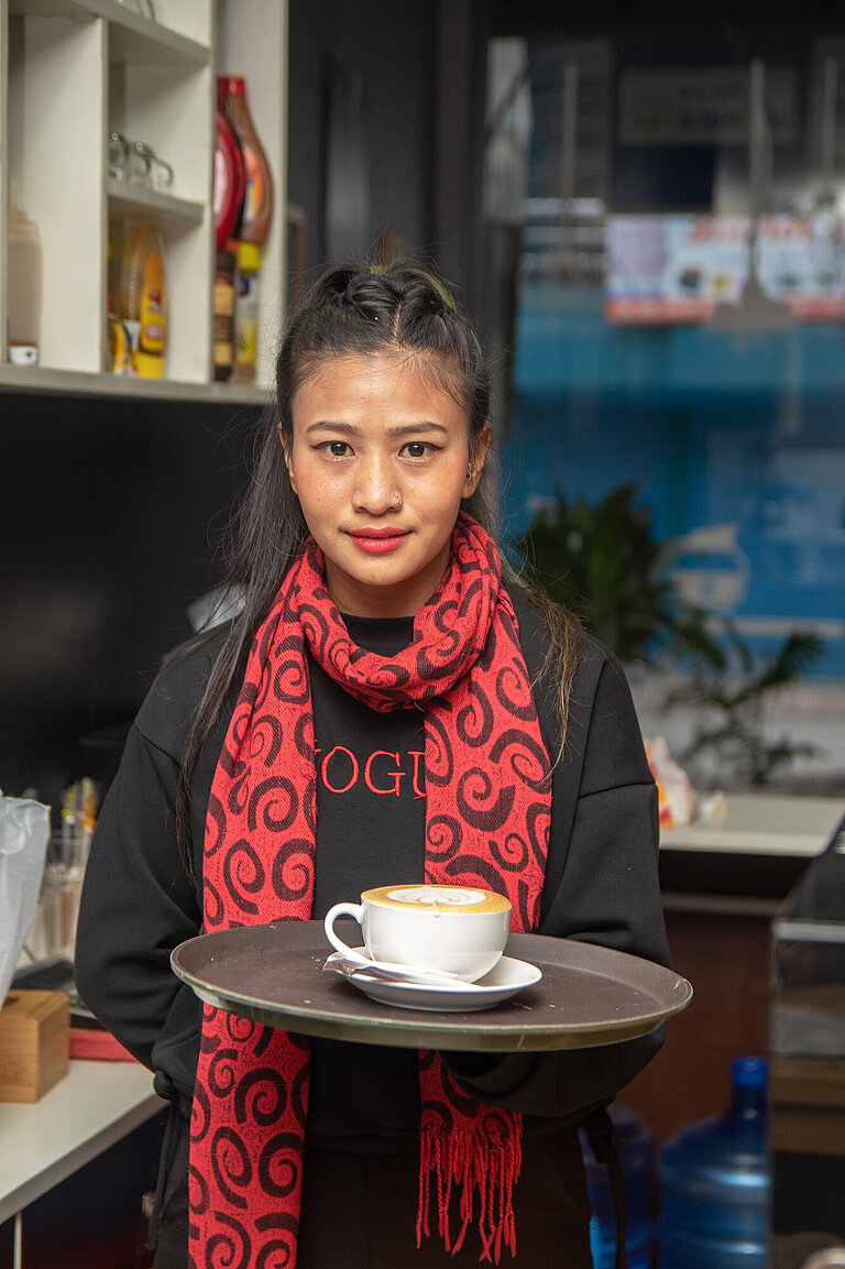 Eine junge Frau trägt einen Schal und hält ein Tablett mit einer Tasse Milchcafé 
