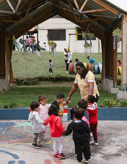 Eine Betreuerin in einer bunten Kleidung steht mit einer Gruppe von Kleinkindern im Kreis unter einer Holzüberdachung. Im Hintergrund sind weitere Kinder auf einem Spielplatz.