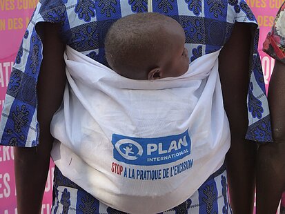  Eine Person trägt ein Baby in einem weißen Tragetuch mit dem Logo von Plan International und dem Schriftzug „Stop à la Pratique de l’Excision“. 