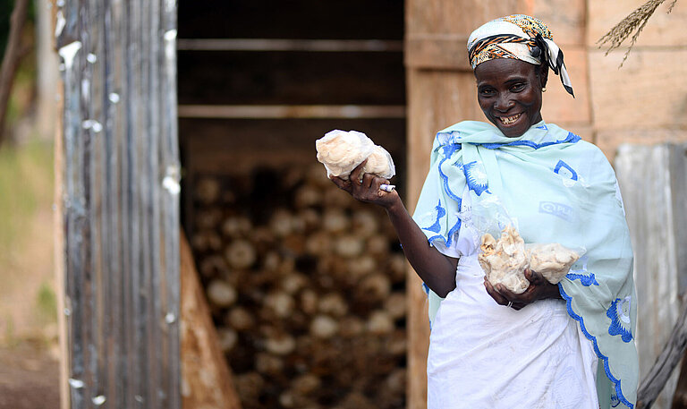Eine Frau steht vor einem kleinen Haus. Sie hält Plastiktüten in der Hand, die voll mit kleinen, hellen Pilzen sind.