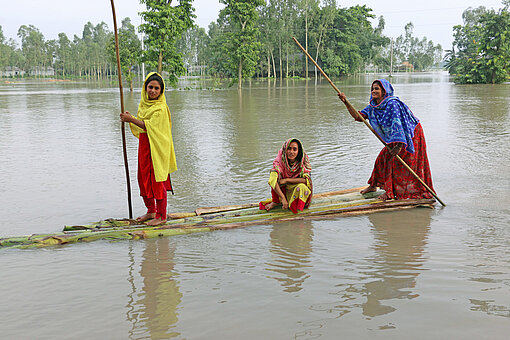 Ein selbst gebautes Floss aus Bambusstangen mit drei Frauen darauf durchfährt eine überschwemmte Landschaft mit Bäumen