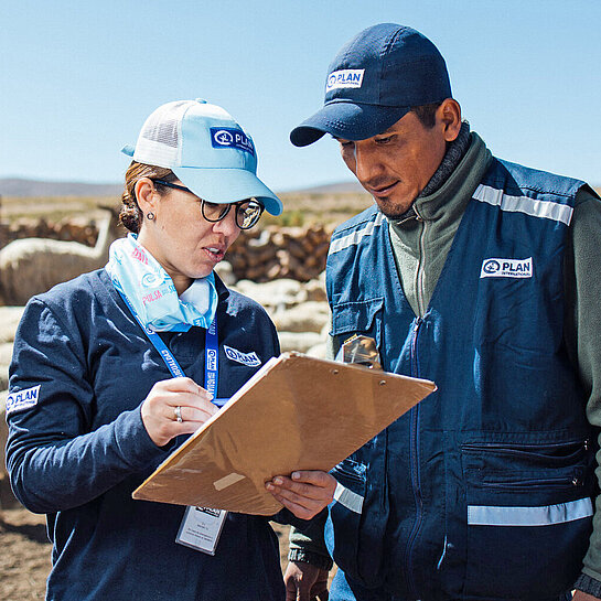 Eine Frau und ein Mann mit dem Logo von Plan International auf ihrer Bekleidung beraten sich über eine Liste gebeugt