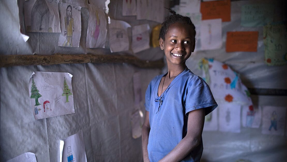 Eyerus lebt mit ihrer Großmutter im IDP Camp in Tigray. Ein Mädchen steht seitlich zur Kamera. Sie schaut über ihre Schulter und Lacht.