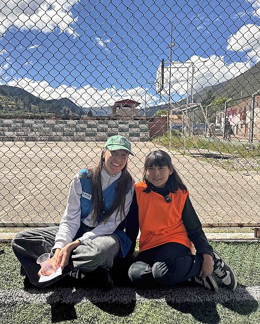 Eine Frau mit blauer Schirmmütze und blauer Weste mit dem Plan-Logo drauf sitzt neben einem kleinen Mädchen am Rande eines Fußballplatzes. Im Hintergrund sind die Berge der Anden in Peru zu sehen.