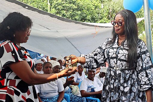 Symbolische Schlüsselübergabe: Miriam Murray, Länderdirektorin von Plan International Liberia (l.), übergibt die „Friedenshütte“ an die Gemeinde von Todee Zwei Frauen in bunten Kleidern reichen sich feierlich ein Schlüsselbund