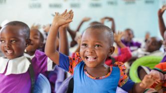 Eine große Gruppe von jungen Kindern sitzt in bunten Kleidern in einer Schule in Tansania. Viele haben die Arme freudig in die Luft gestreckt und lachen herzhaft.