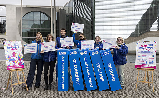 Mitarbeiterinnen von Plan International bei der Domino-Aktion in Berlin Eine Gruppe Menschen steht hinter übergroßen blauen Dominosteinen aus Schaumstoff, auf denen Botschaften rund um das Thema Gleichberechtigung zu lesen sind