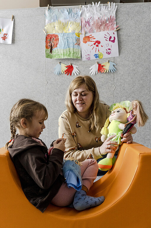 Eine Frau mit einer Puppe in der Hand spricht mit einem kleinen Mädchen.