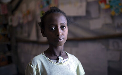 Ein Mädchen in grünem T-Shirt schaut ernst in die Kamera. Hinter ihr hängen bunte Bilder an der Wand.