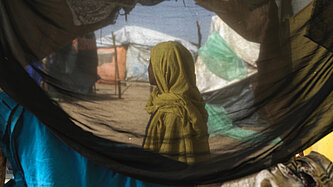 Eine sudanesische Frau steht in einem Geflüchtetencamp, ihr Gesicht ist nicht zu erkenne. Sie ist durch ein Zelt hindurch fotografiert.