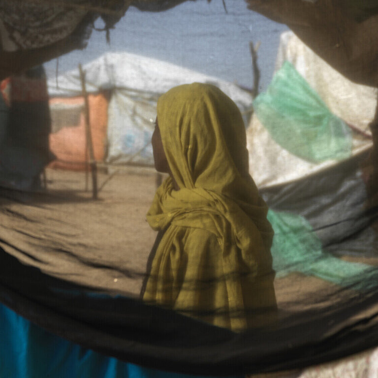 Eine sudanesische Frau steht in einem Geflüchtetencamp, ihr Gesicht ist nicht zu erkenne. Sie ist durch ein Zelt hindurch fotografiert.