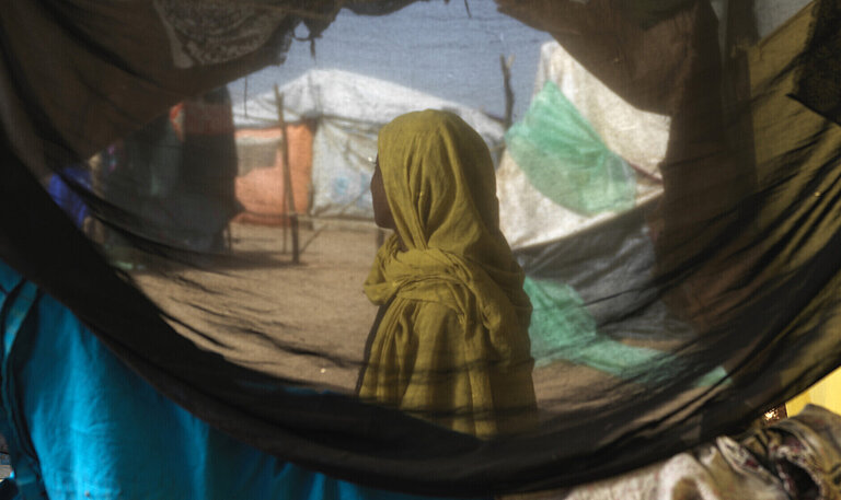 Eine sudanesische Frau steht in einem Geflüchtetencamp, ihr Gesicht ist nicht zu erkenne. Sie ist durch ein Zelt hindurch fotografiert.