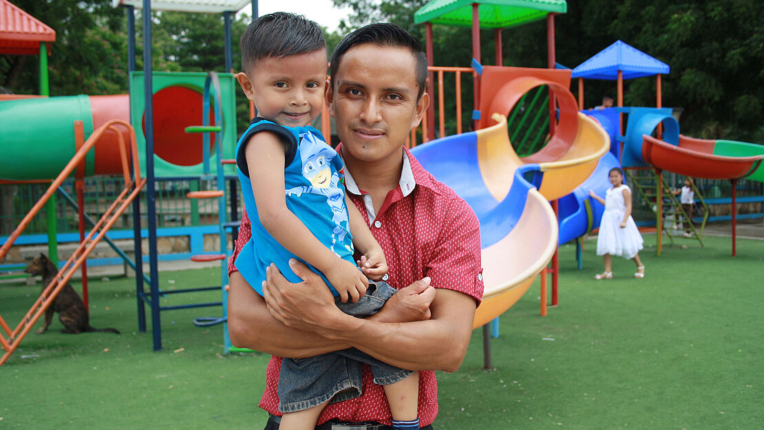 Ein Vater hat seinen Sohn auf dem Arm. Sie stehen auf einem Spielplatz.