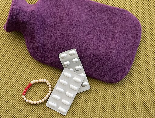 Hilfsmittel gegen Periodenbeschwerden Eine Wärmflasche, zwei Blister Schmerzmittel und ein Armband mit fünf roten und 23 weißen Perlen liegen nebeneinander