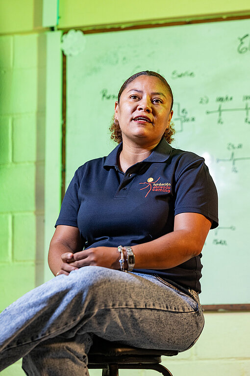 Eine erwachsene Frau sitzt in einem Schulungsraum vor einer Tafel mit elektrischen Schaltplänen und begleitet die technische Ausbildung im Solar‑Girls‑Projekt.