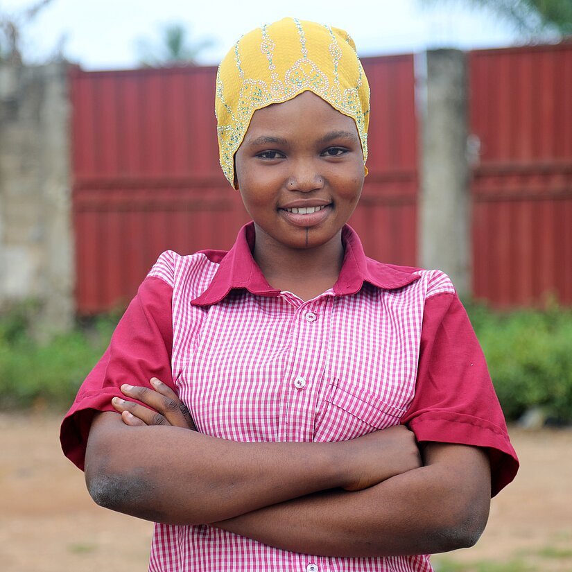 Die 17-jährige Roumana aus Togo trägt ihre Arbeitskleidung. Sie hat ihre Arme vor ihrem Oberkörper verschränkt und lächelt entschlossen in die Kamera.