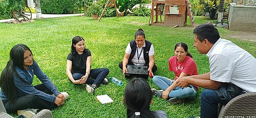 Sechs junge Erwachsene sowie ein Mann sitzen in einem Kreis auf einer saftig grünen Wiese im Garten. Die meisten hören einer Frau aufmerksam zu, die in der Mitte sitzt und eine Mappe in den Händen hält.