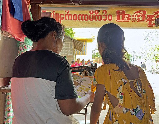Eine Frau und ein Mädchen in bunten Kleidern sitzen unter einer Überdachung
