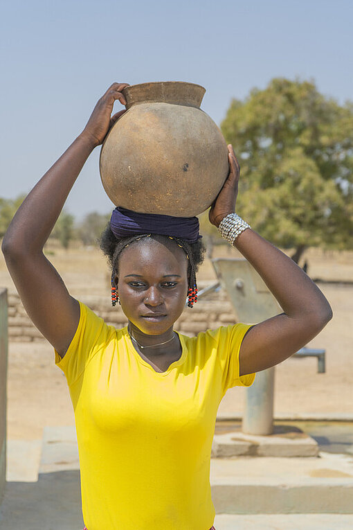 Nassiratou mit Tonkrug voll Wasser auf dem Kopf Mädchen in gelbem T-Shirt trägt Tonkrug aus Wasser auf dem Kopf