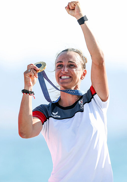 Olympiasiegerin und Plan-Botschafterin Lara Vadlau Eine Frau reckt erfreut die linke Hand in die Luft und hält in der anderen Hand eine golden glänzende Medaille