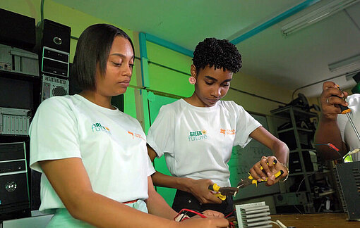 Zwei junge Frauen arbeiten gemeinsam in einem Schulungsraum an elektronischen Geräten; sie tragen Projekt‑T‑Shirts und nutzen Werkzeuge, während im Hintergrund Regale mit Technikmaterial zu sehen sind.