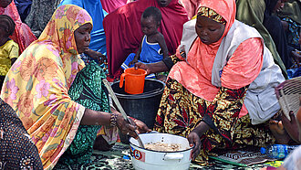 Zwei Frauen sitzen vor einem Topf und kochen, im Hintergrund sitzen weitere Frauen im Schatten eines Baumes