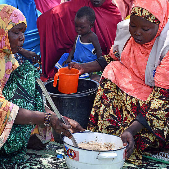 Zwei Frauen sitzen vor einem Topf und kochen, im Hintergrund sitzen weitere Frauen im Schatten eines Baumes