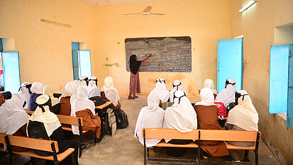 Eine Lehrerin steht vor einer Tafel und unterrichtet eine Klasse von Schülerinnen, die in einem einfachen Klassenraum auf Holzbänken sitzen. Die Schülerinnen tragen helle Kopftücher und Schuluniformen, während die Lehrerin auf die beschriebene Tafel zeigt.