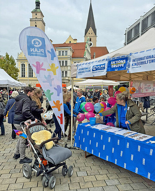 Blick auf einen Marktplatz zwischen historischen Gebäuden und Kirchtürmen, davor ein Info-Tisch mit Personen und dem Plan-Logo