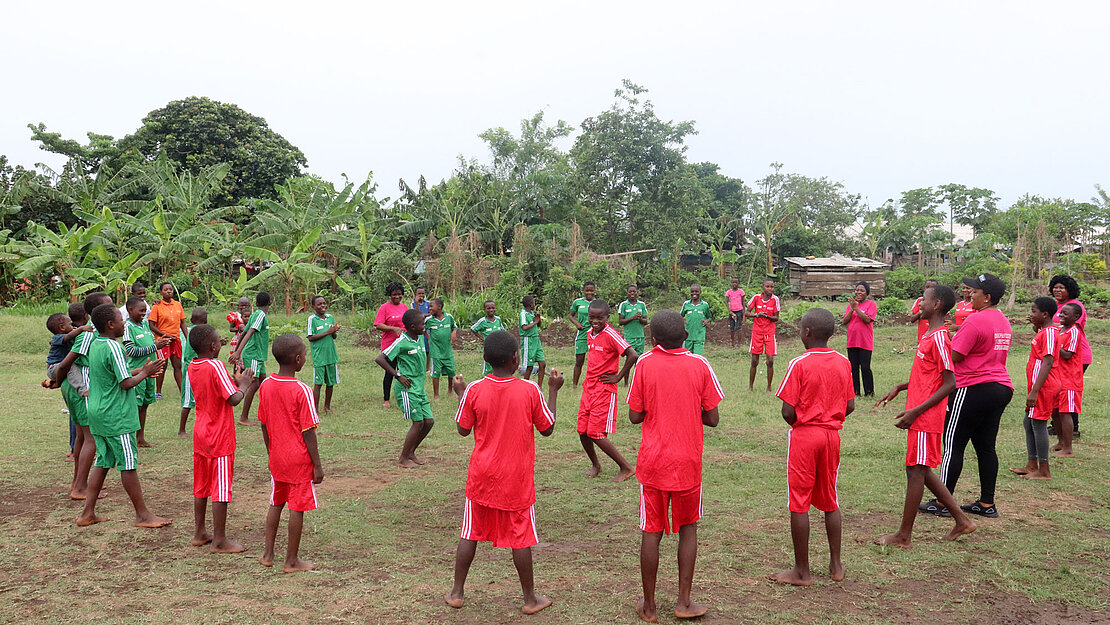 Eine große Gruppe Kinder in Fußballtrikots steht in einem weiten Kreis zusammen, in der Mitte stehen zwei Kinder und tanzen.