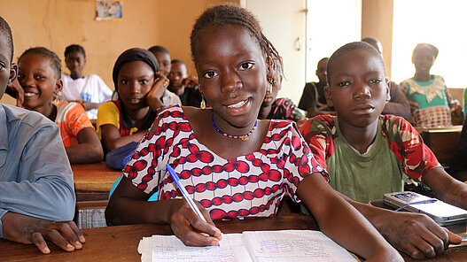 Ein Mädchen schreibt im Unterricht in ihr Heft und lächelt in die Kamera.