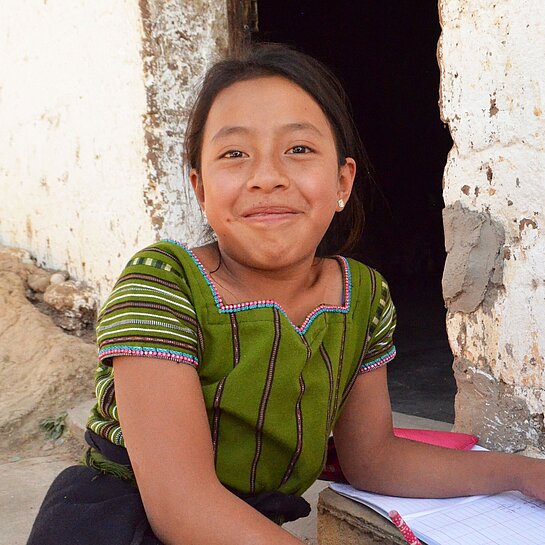 Jenifer mit den Schulsachen, die ihr Plan International gesponsert hat Ein junges Mädchen mit Schulsachen lächelt in die Kamera