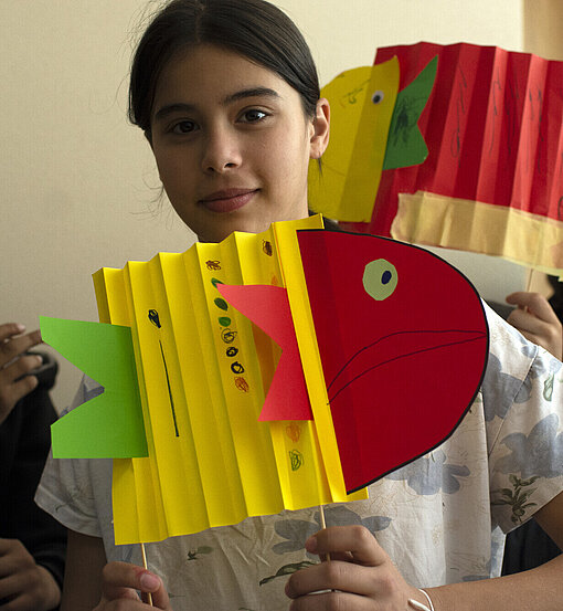 Kunsttherapie Ein Mädchen zeigt einen bunten Fisch aus Papier, den sie gebastelt hat