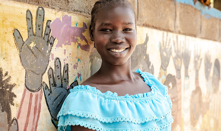 Lächelndes Mädchen in hellblauem Kleid steht mit verschränkten Armen vor einer bunt bemalten Wand mit Handabdrücken.