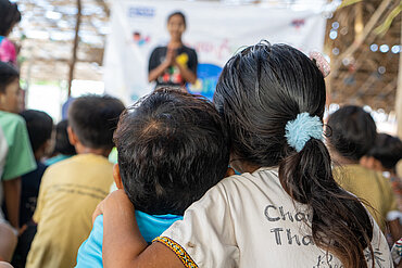 Zwei Kinder, ein Mädchen und ein Junge, sitzen dicht nebeneinander inmitten einer größeren Gruppe und haben die Arme umeinander gelegt. Das Mädchen trägt einen Zopf mit einem blauen Haargummi. Beide blicken aufmerksam nach vorne, wo eine Betreuerin auf einer Bühne steht und spricht.