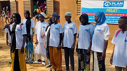Eine Gruppe von jungen Männern und Frauen in traditioneller Kleidung und weißen T-Shirts von Plan International steht vor einem Gebäude. Eine Person spricht in ein Mikrofon, während weitere Personen und Kinder im Hintergrund zuschauen. 